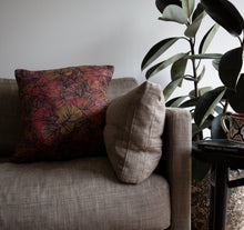 Load image into Gallery viewer, Our organic cotton cushion cover with repeated fan palm leaf pattern in orange, red, purple, mauve and yellow colours with a black outline. The cushion is sitting on a couch and next to it is a small table with Leif Cocks book 'Finding Our Humanity' and a mug, and a plant in the background.