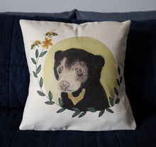 Load image into Gallery viewer, Sitting on a bed is our organic cotton cushion cover with an original watercolour and hand stitched illustration of a sun bear cub, a pale yellow circle sun behind the bear, leaves framing the image with a yellow flower at the end and two bees.