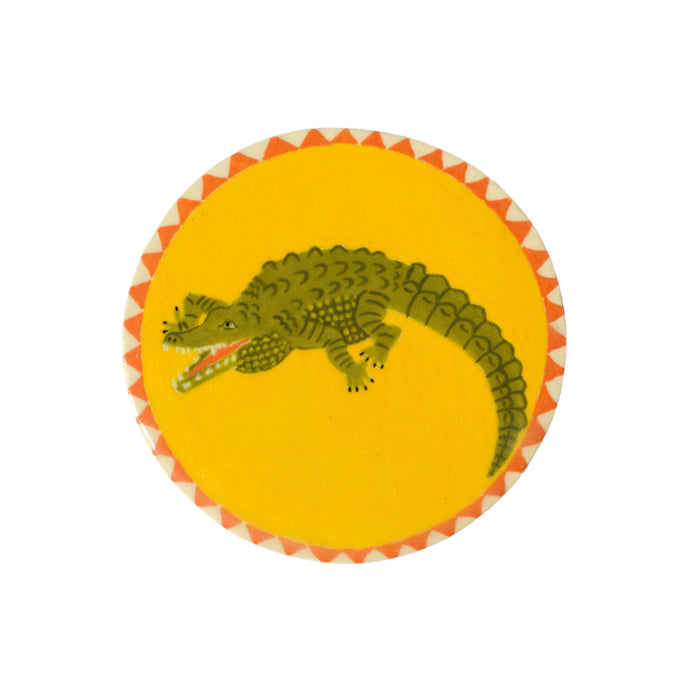 A circular ceramic tile with a green crocodile, bright yellow background and an orange and white border of triangles