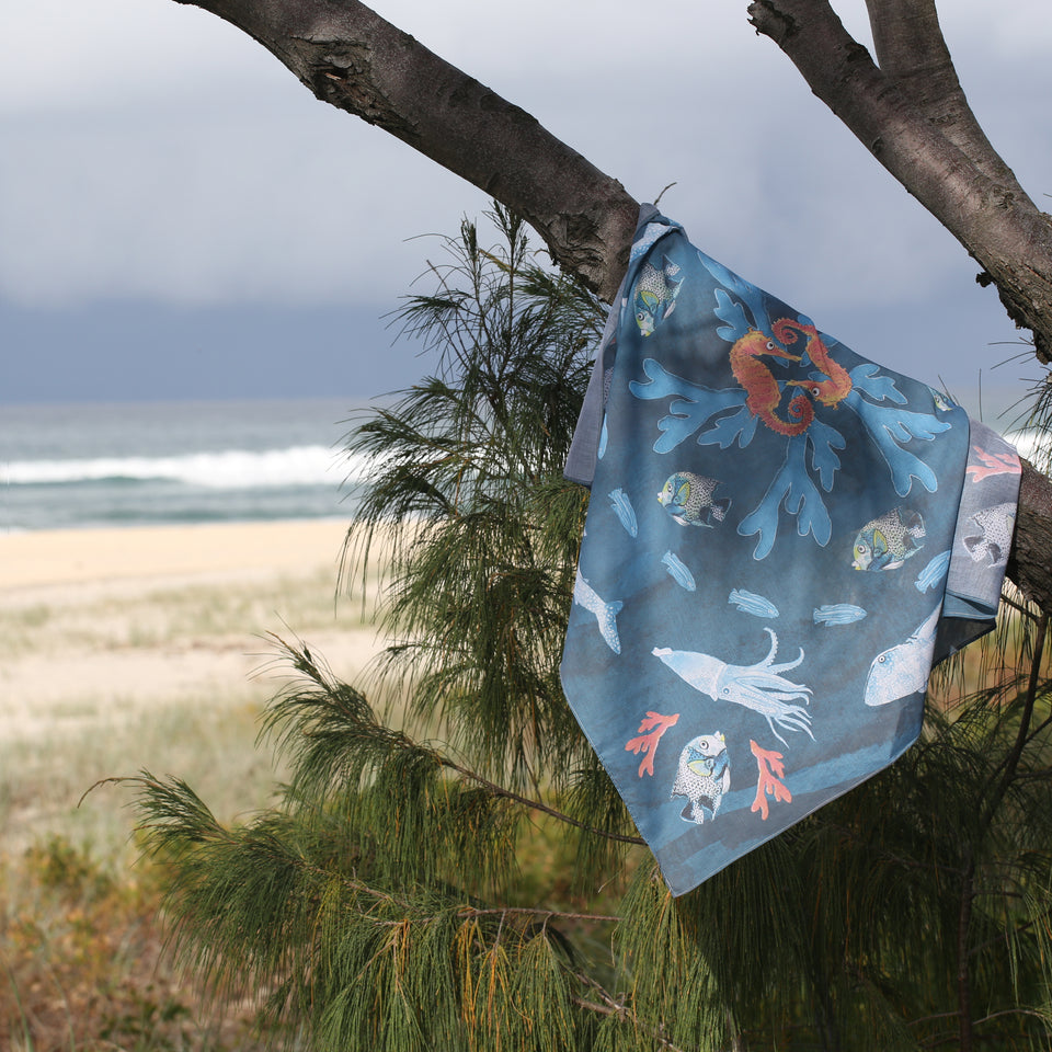 Nyattski Sea Life scarf draped over a tree branch at the beach with the sand and ocean showing in the background. 