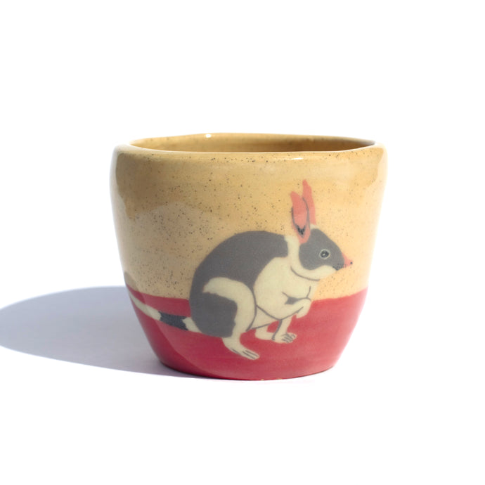 A  handmade ceramic cup in a light coloured speckled clay with a hand drawn illustration of a bilby and a red colour along the bottom like the desert sand. 