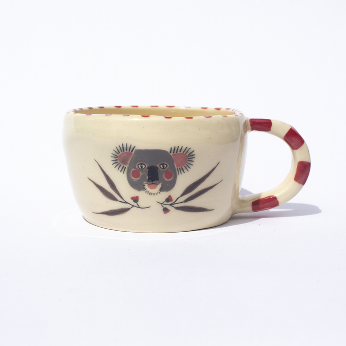 Handmade ceramic mug with a pink striped handle and illustrations of a koala face with pink rosy cheeks, gum leaves and gum flowers.