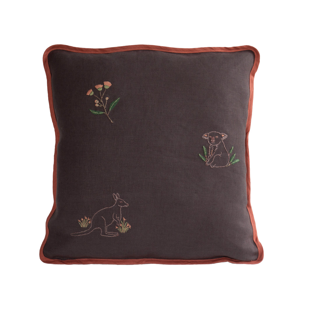 Hand embroidered pink flowering gum, a sitting koala with some leaves, and a kangaroo with kangaroo grass next to it, on a dark grey linen cushion with vintage pink coloured binding.