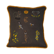 Load image into Gallery viewer, Grey coloured cushion with mustard yellow binding. Embroidered on the cushion is a brushtail possum, sun, half moon with a face, koala face with wattle underneath, red wattle bird, bee, wattle, wallaby and more wattle.
