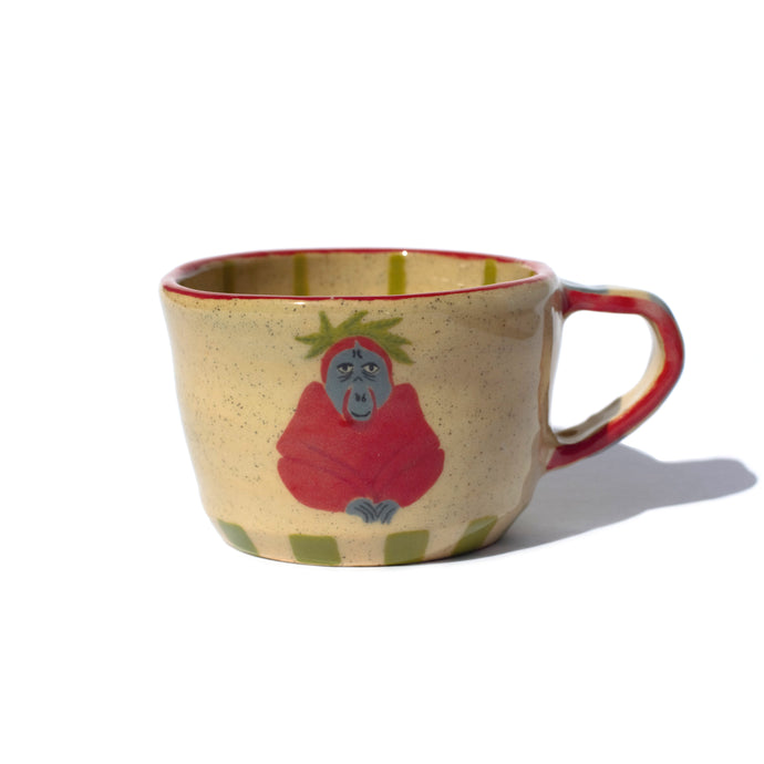 A handmade ceramic mug with an illustration of a sitting orangutan with leaves on its head, green colour squares along the bottom, side of handle and rim painted red, and thin green stripes on the inside. 