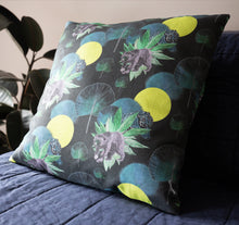 Load image into Gallery viewer, Organic cotton cushion with a repeat pattern of a sun bear, a blue tiger, yellow circle sun, green and blue fan palm leaves and plants  on a dark coloured background. The cushion is sitting on a bed with a plant in the left hand corner.
